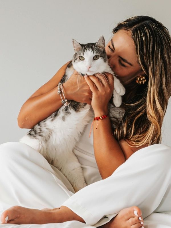 A person in white clothing is sitting on a white bed, holding and kissing a grey and white cat.