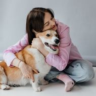 A person in a pink shirt hugs a corgi dog.