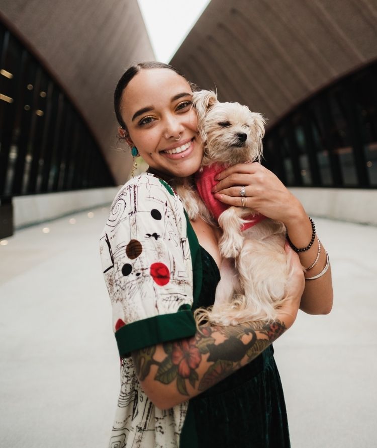 A person holds a small, light-colored dog wearing a pink shirt. The person has tattoos on their arms and is smiling at the camera. They are standing outside with a modern building in the background.