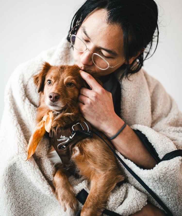 A person wearing glasses and a fluffy robe gently kisses a small brown dog wearing a harness.